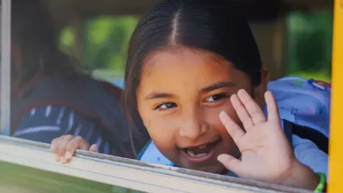 Young female student waving her hand outside a bus window.