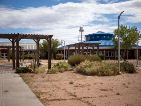 School courtyard with wooden pergolas, desert landscaping, a blue-roofed building, and a water tower under a partly cloudy sky.