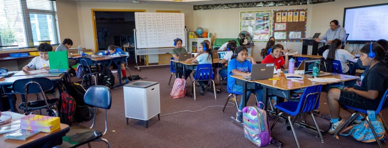 Students work independently and in small groups on laptops and assignments while a teacher instructs at the front of the classroom.