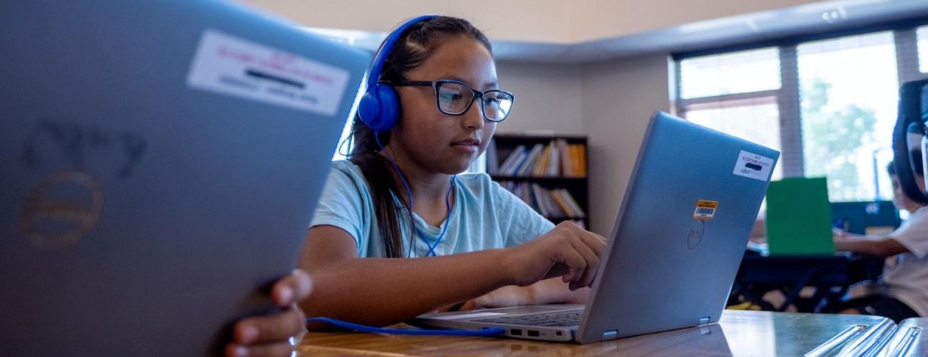 Student wearing glasses and blue headphones works on a laptop in a classroom computer lab.